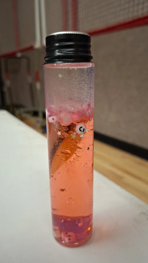 Tall pink liquid-filled bottle with floating objects on a white table.