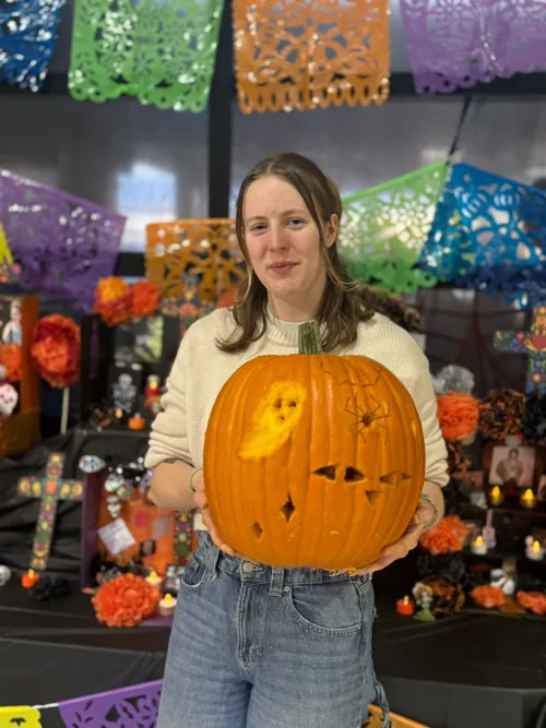 Person holding a carved pumpkin with a ghost design, surrounded by colorful decorations.