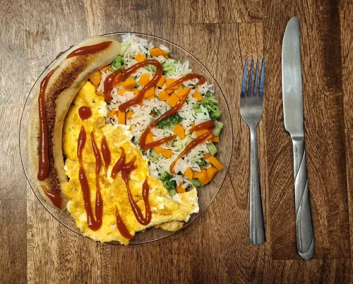 Omelet with sauce, rice, vegetables, and a banana on a plate; fork and knife on the side.