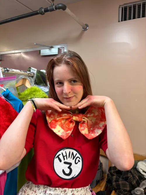 Young woman in a red shirt with a large floral bow tie, smiling and posing with hands under chin.