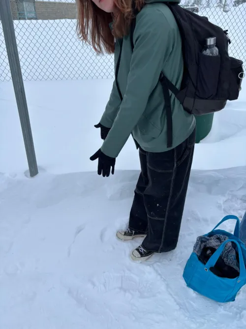 Person in a green jacket and gloves pointing at snow, with a blue bag nearby.