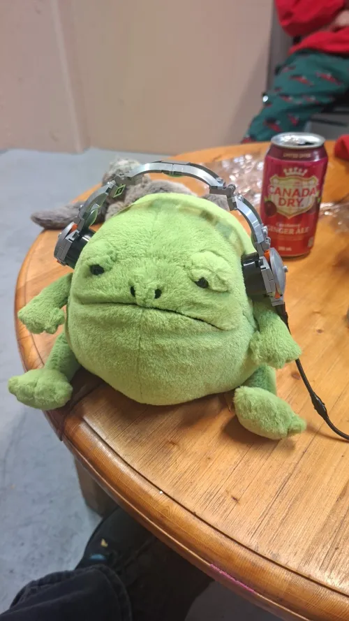 Green frog plush with headphones on a wooden table.