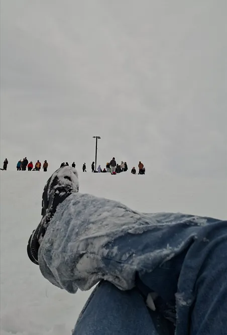 Snow-covered legs in jeans, people on hilltop in distance.