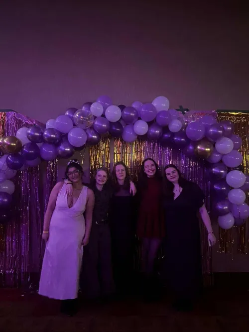 5 people smiling under a purple balloon arch at a party.