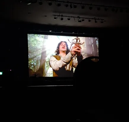 Movie screen showing a person holding a sword in a forest scene.