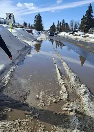 Snowy street with slushy puddles and a distant car; clear blue sky overhead.