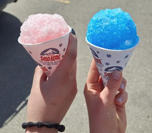 Two hands holding pink and blue snow cones under sunlight.